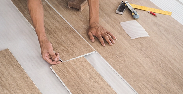 A person measuring and installing light wood laminate flooring with tools nearby.