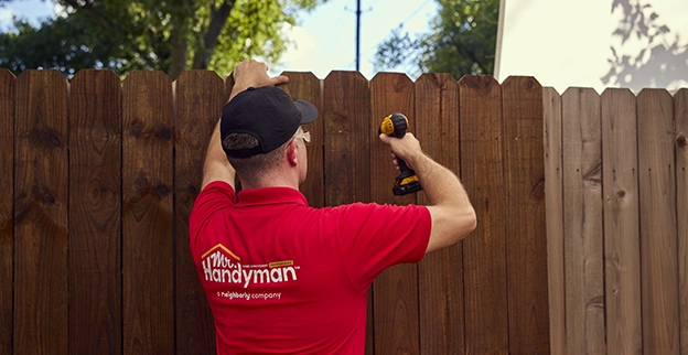 A handyman in a red shirt uses a power drill to repair a wooden fence outdoors, focusing on the task at hand.