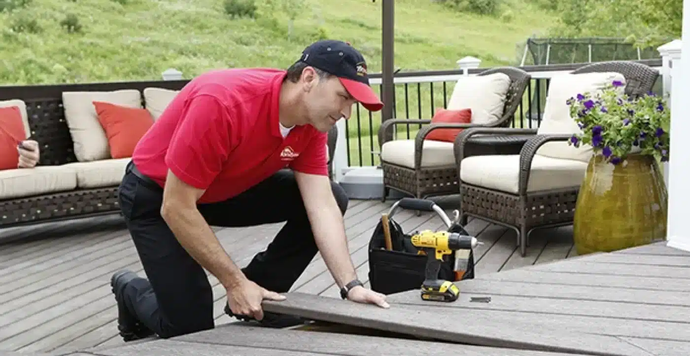 A uniformed Mr. Handyman service professional kneeling to lift and replace a deck board during an outdoor deck repair project.